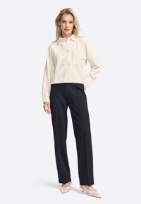 Crème cropped button-up jasje met twee voorzakken, gecombineerd met zwarte op maat gemaakte broeken. Het model draagt beige puntige hakken.
