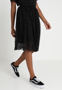 Musta, läpikuultava polkadot-mittainen hame, jossa on joustava vyötärönauha, yhdistettynä mustiin lenkkareihin. Kevyt ja liehuva design. Havainnoitu liikkeessä.