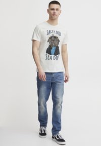 Lys creme bomulds T-shirt med en illustration af en mops i kaptajnens outfit. Sammen med blå denim jeans og sorte sneakers.