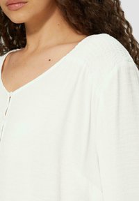 Witte, gestructureerde blouse met een V-hals, voorzien van een knoopsluiting en geplooide schouderdetails. Zachte stof, losse pasvorm.