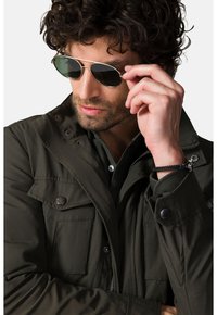 Oliwkowa kurtka z wieloma kieszeniami, z guzikami i kapturem. Posiada srebrne okulary przeciwsłoneczne oraz czarną teksturowaną bransoletkę na nadgarstek.