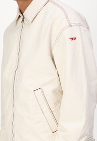 Chaqueta de algodón beige con cremallera, bolsillo de parche, costuras rojas en contraste y un pequeño logo rojo en la manga.
