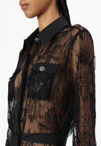 Blusa nera in pizzo con una texture trasparente, dotata di colletto a punta, taschini sul petto con bottone e hardware decorativo.