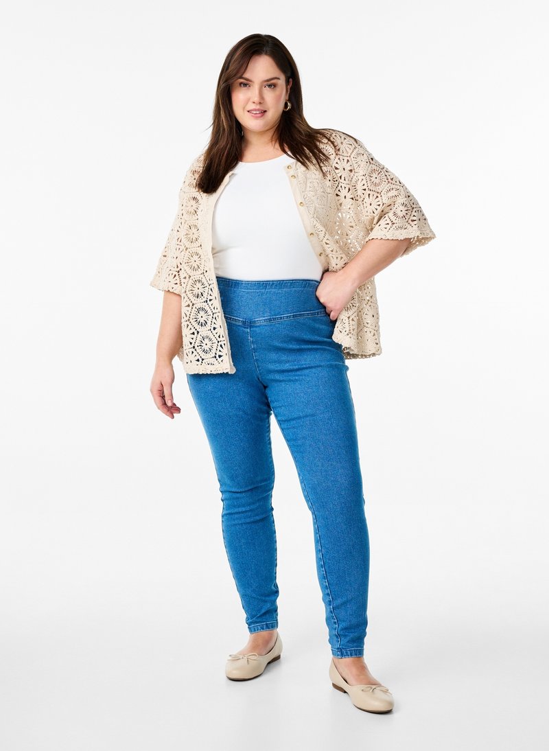 Žena nosiaca béžový háčkovaný kardigán cez biely top, modré nohavice skinny s vysokým pásom a béžové balerínky, stojaca s jednou rukou na bok.
