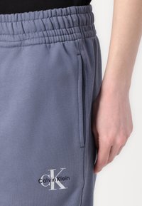 Sive pamučne trenirke s elastičnom uvlačnom trakom, bočnim džepom i bijelim Calvin Klein logom vezanim na lijevom bedru.
