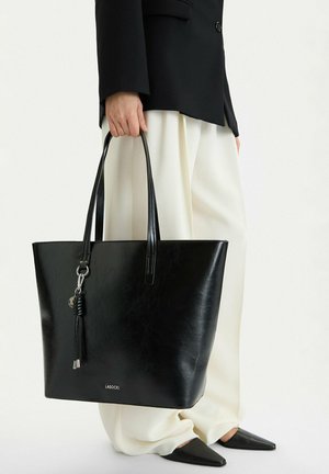 Persona sosteniendo un gran bolso tote de cuero negro con detalle de flecos, vistiendo un blazer negro, pantalones anchos blancos y zapatos negros puntiagudos.