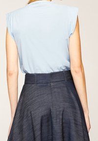 Donna che indossa un top senza maniche di colore azzurro chiaro, infilato in una gonna A-line di jeans scuri a vita alta con passanti per la cintura, fotografata di spalle.