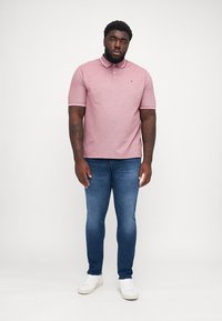 Uomo adulto in piedi, che indossa una polo rosa a maniche corte, jeans blu e sneaker bianche, su uno sfondo chiaro e semplice.