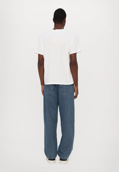 Homme debout, vu de dos, portant un t-shirt blanc uni à manches courtes, un jean bleu ample avec des poches arrière, et des baskets blanches.