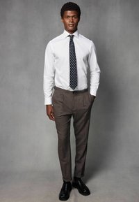 Hvid skjorte med knap, marineblå slips med prikker og brune, strukturerede bukser. Sorte sko fuldender det skræddersyede look. Modellen står oprejst.