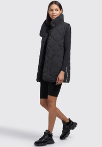 Gilet sans manches matelassé noir avec un grand motif en relief, col montant, fermetures à pression et poches latérales, associé à un short noir et des chaussures.