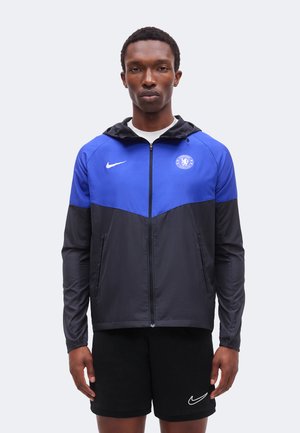 Mand iført en blå og sort Chelsea FC lynlåstrøje med hætte og sorte Nike-shorts, stående mod en ensfarvet lys baggrund.