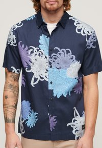 Camisa de manga corta hecha de tela con fondo azul marino, que presenta grandes patrones florales en azul claro, lavanda y blanco. Botones negros visibles.