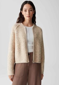 Beiger, flauschiger Cardigan mit Reißverschluss und rundem Halsausschnitt. Verfügt über gerippte Akzente an den Bündchen und am Saum, kombiniert mit einem weißen Oberteil darunter.