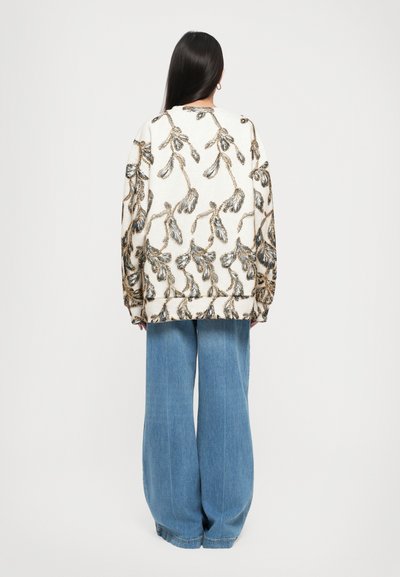 Woman with long dark hair wears white oversized sweater with black and gold leaf pattern and wide-leg blue jeans, facing away.