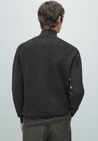 Zwarte gebreide trui met een hoge kraag, een gestructureerde stof en geribbelde details aan de manchetten en zoom, gedragen over een grijze laag.