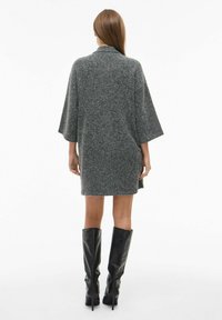 Pilkos vilnos mišinio per didelis suknelė su plačiomis rankovėmis, turinti tiesų siluetą ir kontrastingais iki kelių juodais odiniais batais.