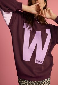 Bordeauxfärgad sweatshirt med en stor lila "W"-logotyp, med kontrasterande ljusrosa ärmar och en avslappnad passform. Bärs med leopardmönstrade byxor.