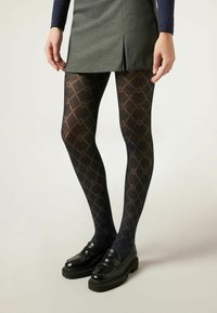 Des collants noirs à motif avec un design en diamants et des accents marron clair, associés à des mocassins noirs et une jupe grise.