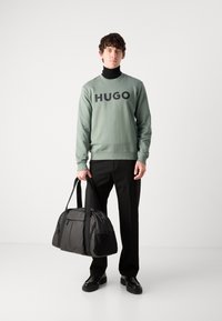 Grønn hettegenser med "HUGO" trykk, svart genser med høy hals, svarte bukser, holder en svart duffelbag med teksturerte aksenter. Svarte sko.