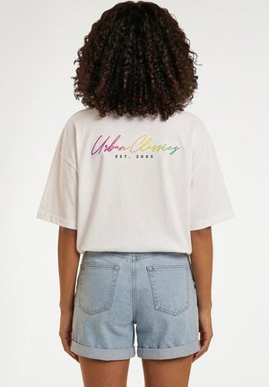Personne aux cheveux bouclés porte un t-shirt blanc oversize avec le texte multicolore « Urban Classics EST. 2005 », associé à un short en jean bleu clair.