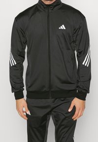 Sort atletisk jakke med høj krave, præget af hvidt Adidas-logo og tre-stribet design på ærmerne, lavet af glat, let materiale.