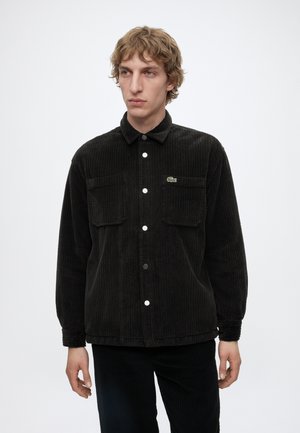 Joven con camisa negra de pana abotonada con bolsillos delanteros y parche con logo, posando frente a un fondo blanco liso.