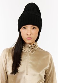 Zwarte geribbelde brei-beanie met een spitse top, gecombineerd met een lichtgouden ritsshirt met een gladde, reflecterende textuur.