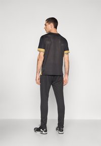 Sort atletisk t-shirt med guldaccenter, korte ærmer og sideslids, parret med sorte tapered bukser og sorte sneakers.
