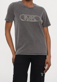 Camiseta gris de manga corta hecha de algodón con cuello redondo y diseño de logotipo con tachuelas en la parte delantera. Textura suave con un ajuste relajado.