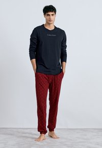 Cămașă neagră cu mânecă lungă, având logo-ul Calvin Klein, asortată cu pantaloni de pijama roșii cu model. Design simplu, material moale, croială relaxată.