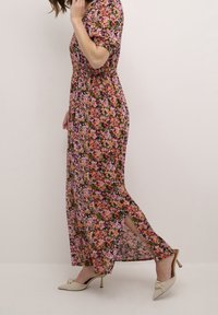 Robe longue fleurie à manches courtes, avec un fond noir orné de fleurs roses, orange et jaunes. Comprend des fentes latérales.