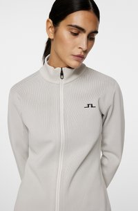 Veste zippée gris clair avec un col montant, dotée de panneaux en maille respirante sur les épaules et d'un petit logo noir sur la poitrine gauche.