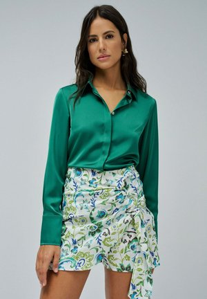 Camisa de satén verde esmeralda con mangas largas y cuello, combinada con una falda envolvente blanca con patrones florales en azul y verde.