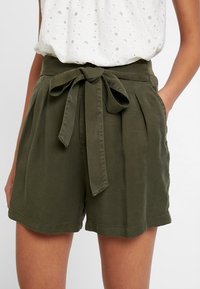 Shorts vert olive en coton doux avec une taille ceinturée, des plis à l'avant et des poches latérales. L'ourlet est fini par un revers simple.