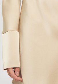 Beige samettitoppi, joka korostaa leveää hihansuun muotoilua, sileää rakennetta ja hienovaraista kiiltoa. Läheltä otettu kuva kankaasta ja käden vuorovaikutuksesta.