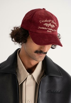 Man with mustache wears maroon cap with floral embroidery and text, beige buttoned shirt, and black leather jacket with zipper and embroidered text.