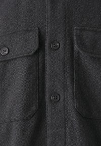 Czarna, teksturowana koszula z dwiema kieszeniami na piersi, zapięciem na guziki oraz kontrastującymi czarnymi guzikami, z delikatnym geometrycznym wzorem.