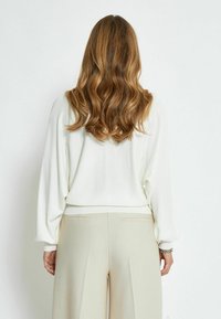 Maglione bianco a maglia con maniche lunghe e ampie e orlo a coste, abbinato a pantaloni beige a vita alta con pieghe. Tessuto morbido e testurizzato.