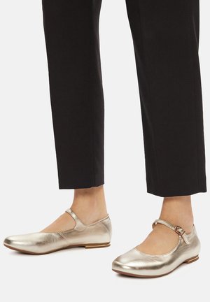 Metaliczne złote baletki z pojedynczym paskiem wokół kostki, zaokrąglonym noskiem i płaską podeszwą, zestawione z czarnymi, dopasowanymi spodniami. Gładka faktura.