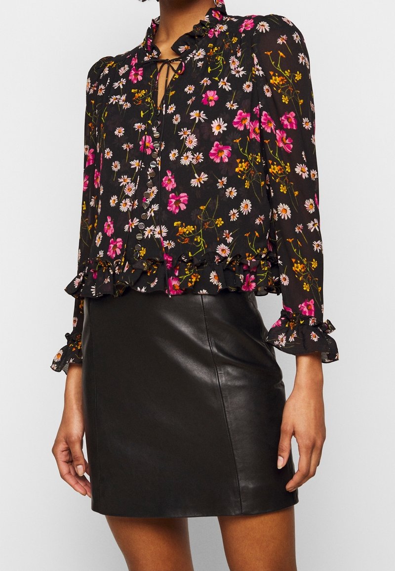 Blouse noire à fleurs avec des fleurs roses et blanches, poignets et ourlet à volants, encolure nouée, associée à une jupe en cuir noire.