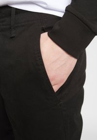 Zwarte broek met een gladde textuur en een elastische tailleband. Eén hand rustende in het zijzakje, waarbij de opening van het zakje duidelijk zichtbaar is.