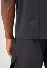 T-shirt de sport noir à manches courtes, texture lisse et coupe ajustée. Coutures visibles le long de l'ourlet et du col.