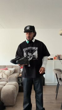 Black athletic shoes with a textured surface, held by a person wearing a black graphic t-shirt and baggy pants, indoors with furniture in background.