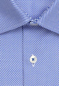 Nærbillede af en skjortekrave med blå og hvide zigzag-mønstre, med en hvid knap fastgjort under kraven.