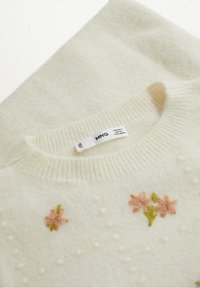 Pull couleur crème avec détail en relief bouclé et broderie florale rose. Encolure côtelée et étiquette visible près du col.