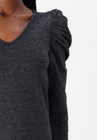 Black, glittery top featuring a deep V-neck, textured fabric, and puffed sleeves, emphasizing a fitted design and subtle shimmer.