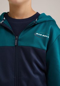 Huvtröja med en blandning av gröna och marinblå paneler, tillverkad av ett smidigt tyg med dragkedja och "BAD BOYS"-logotyp på bröstet.