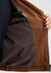 Bruin suède jack interieur met een donkere gladde voering, voorzien van een ritszak en een zachte textuur. Het jack heeft een ritsluiting aan de zijkant.