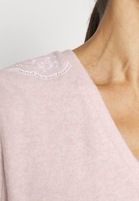 Ljusrosa stickad tröja med V-ringning och subtil vit broderidetalj på axeln. Mjuk textur, närbild.